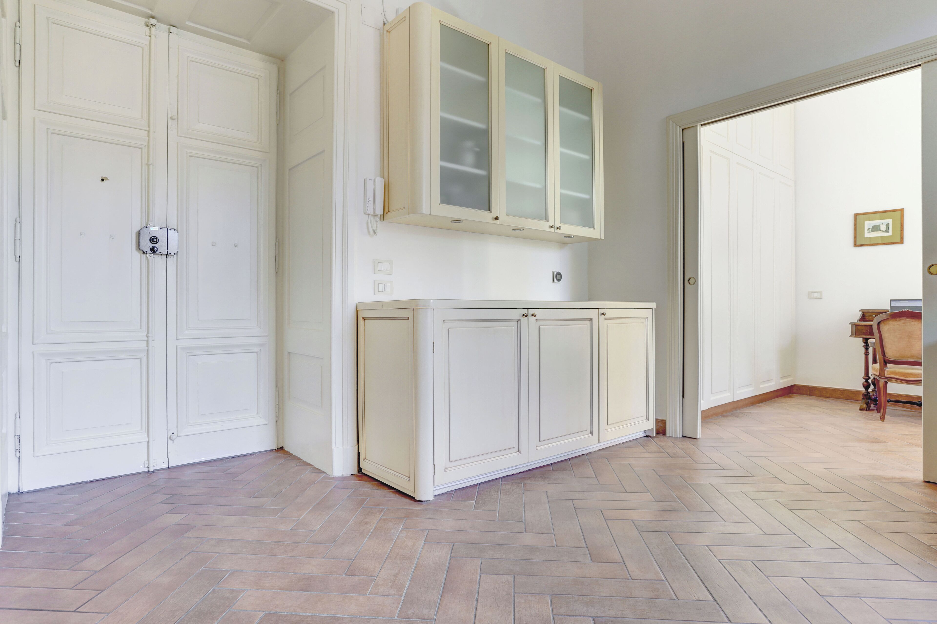 Una habitación con suelo de espiga, puertas dobles blancas, un mueble con puertas de cristal y una silla de madera en la esquina.