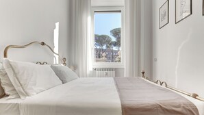 Un llit ben fet amb roba de llit blanca i beix, un reposacapses de fusta, i una finestra amb vista a arbres i edificis.