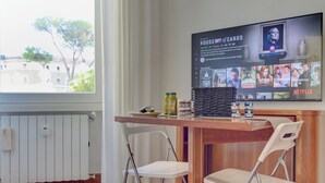 Una habitació amb taula de fusta, dues cadires blanques, un televisor que mostra 'House of Cards' i una finestra amb vista a edificis i arbres.