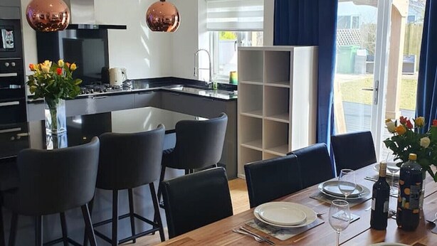 A modern kitchen with a central island, black bar stools, and a dining table set for six with plates and glasses. - Blue Bay Villa in Trearddur Anglesey (Trearddur Bay)