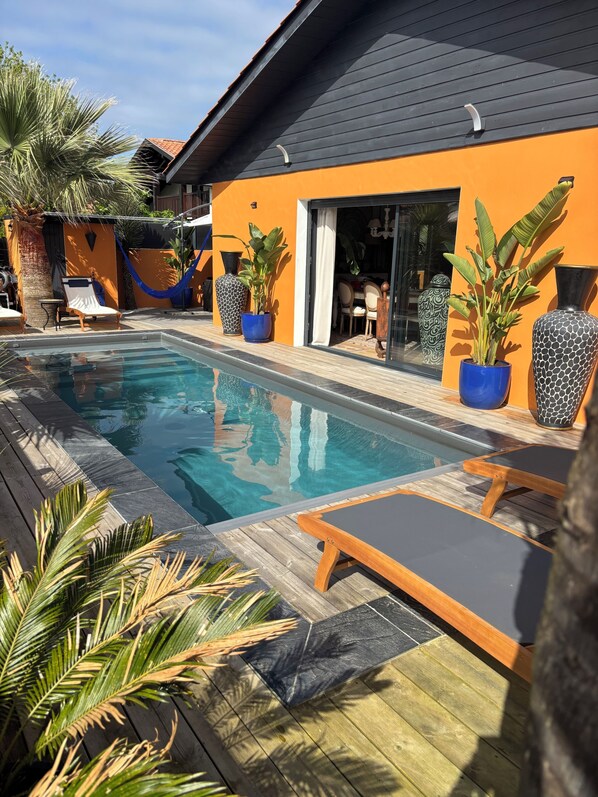 A swimming pool with lounge chairs, potted plants, and a modern building with an orange facade.