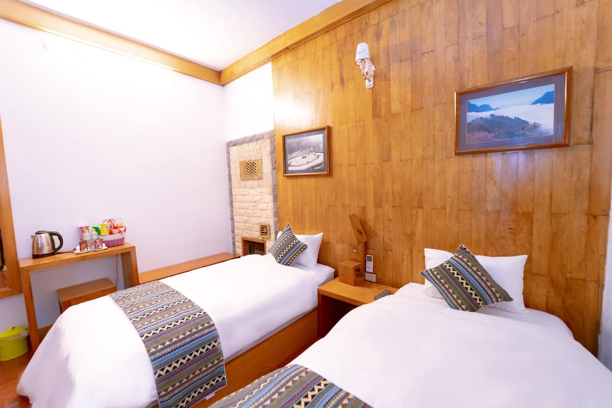 A hotel room with two beds, wooden headboards, a wooden wall, a small table with a kettle and breakfast items, and a framed picture.