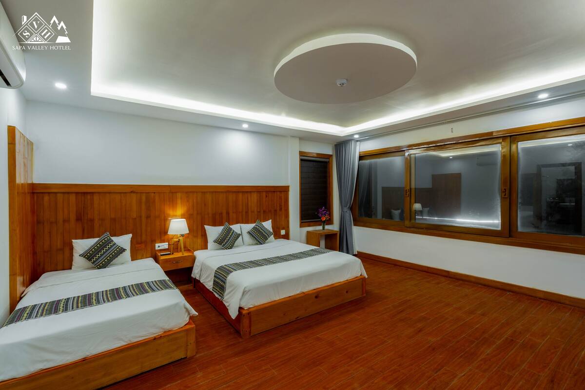 A hotel room with two beds, wooden headboards, a large window with curtains, and a ceiling light fixture.
