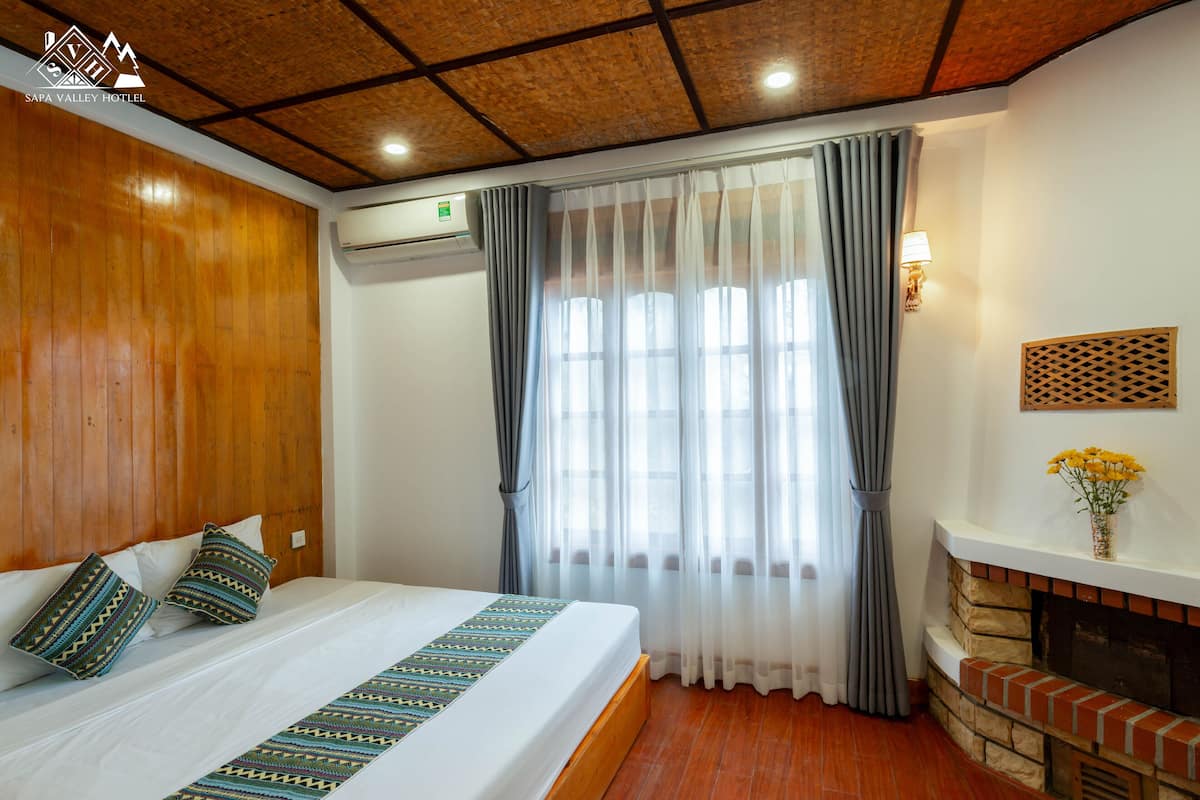 A hotel room with a wooden ceiling, a bed with patterned pillows, a fireplace, and a window with curtains.