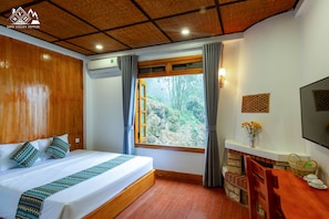 A hotel room with a wooden ceiling, a large bed with patterned pillows, a window with a view of greenery, a small table with a vase and a basket, and a TV mounted on the wall. - Sapa Valley Hotel (Sa Pa)