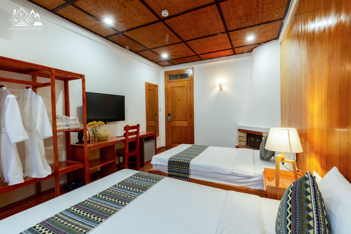 A hotel room with two beds, a wooden wardrobe, a desk with a chair, a television, and a lamp.