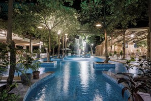 An indoor swimming pool with tiled edges, surrounded by trees and potted plants, and illuminated by overhead lights.