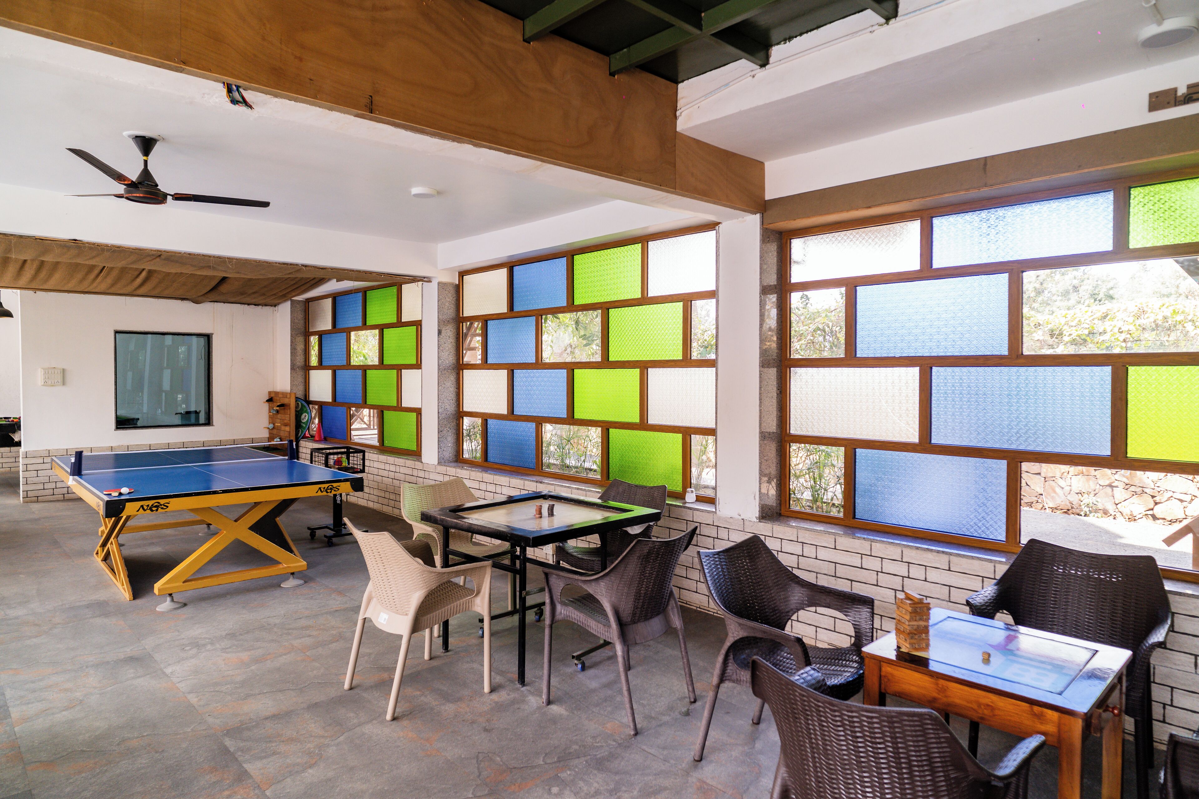 A room with a ping pong table, a pool table, wicker chairs, and large windows with colorful panels.