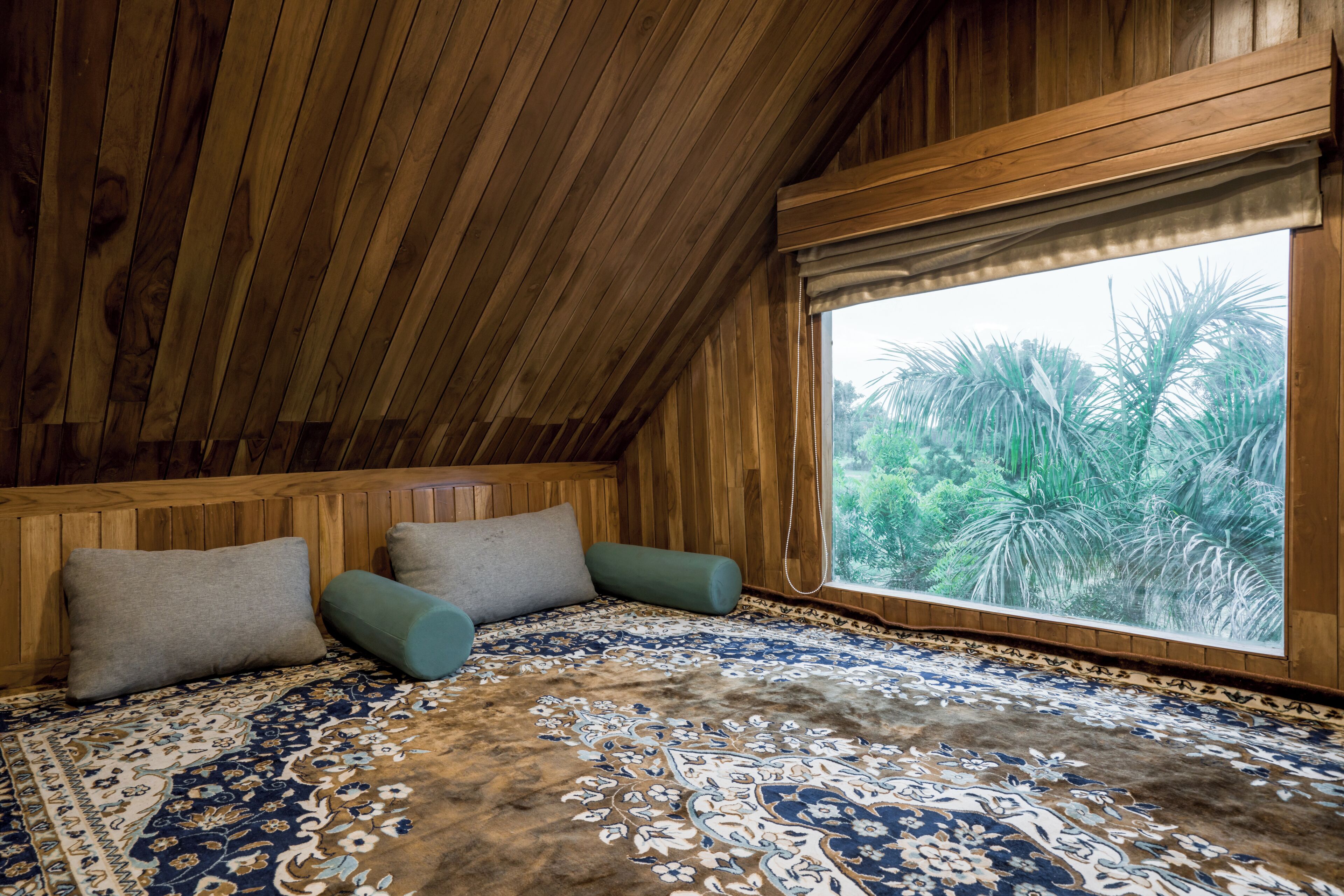 A room with wooden walls and ceiling, a large window with a view of palm trees, a patterned rug, and two pillows.
