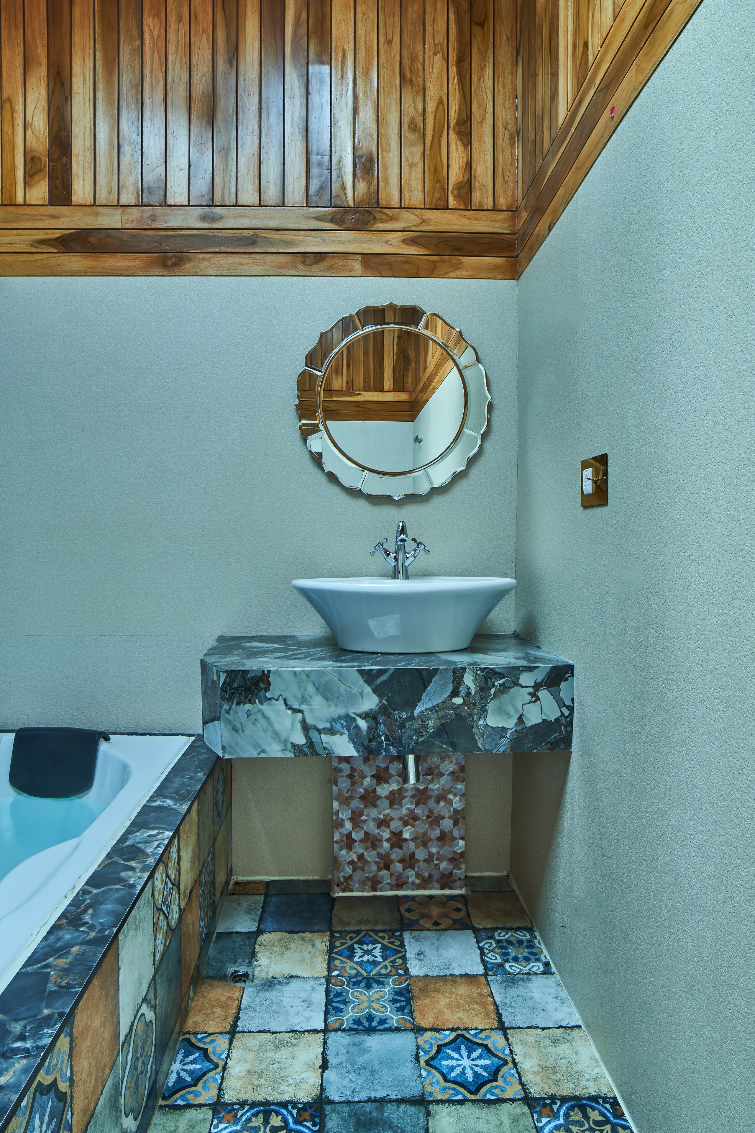 Un baño con techo de madera, un lavamanos con encimera de mármol, un espejo decorativo y un piso de baldosas con diseño geométrico.