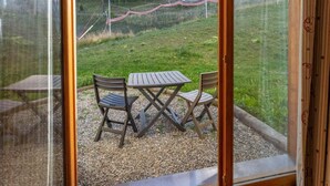 Un patio con mesa y sillas, suelo de grava y vista a un área con césped, árboles y un telesilla al fondo.