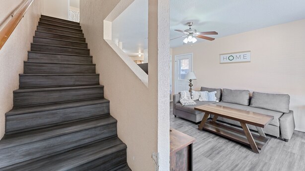 Eine Treppe mit dunklen Stufen führt zu einer Zwischenebene, dort befindet sich ein Deckenventilator, ein Sofa und ein Couchtisch in einem Wohnbereich.
