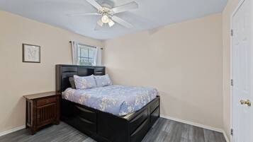 Une chambre à coucher avec un lit, une table de chevet, un ventilateur de plafond et une fenêtre avec des rideaux.
