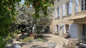Ein Innenhof mit Tisch und Stühlen, umgeben von Pflanzen und einem Gebäude mit Fenstern und Fensterläden.