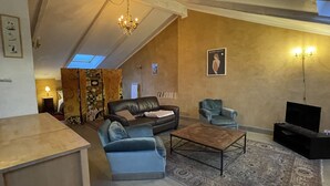 Ein Wohnzimmer mit Sofa, Sessel, Couchtisch und Fernseher. Eine Kandelaber, ein Teppich mit Muster und ein Gemälde an der Wand sind ebenfalls zu sehen.