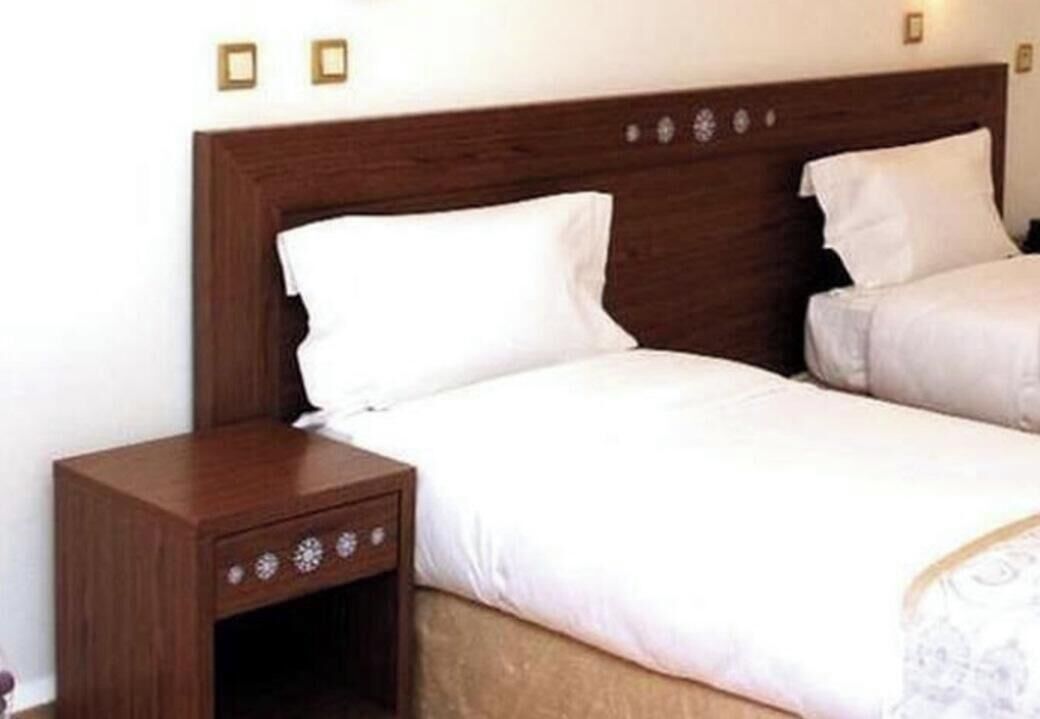 Une chambre à coucher avec une tête de lit en bois, un lit simple avec une literie blanche et une table de chevet dotée d’un tiroir.