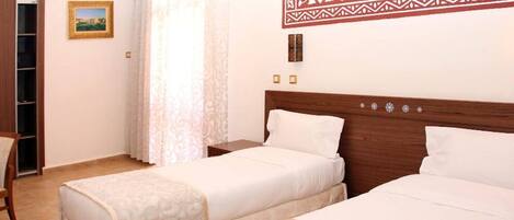 Quarto de hotel com duas camas, cabeceira de madeira, uma mesa pequena, uma cadeira e um guarda-roupa.