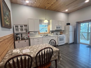 A kitchen with white cabinets, a dining table with a floral tablecloth, and a window with a view of trees.