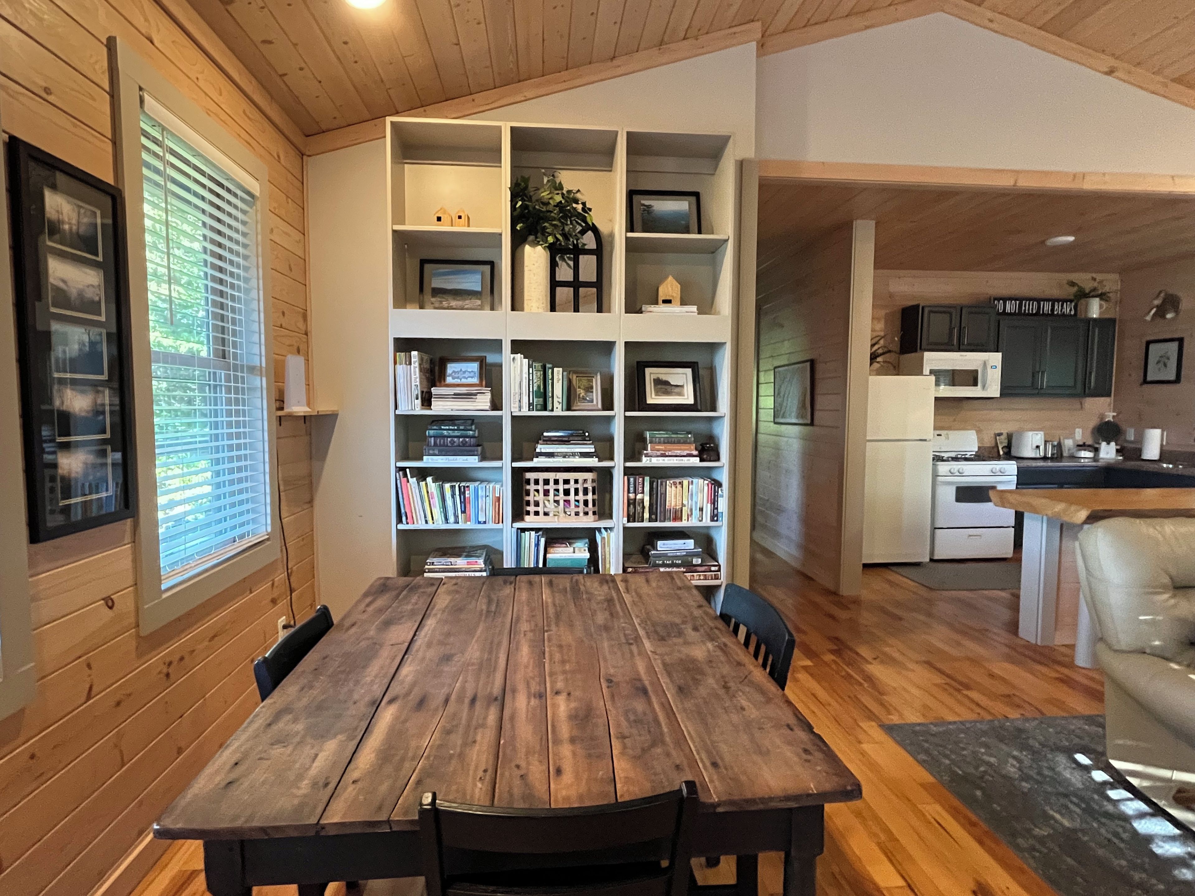 A wooden dining table with chairs, a built-in bookshelf, and a kitchen area with cabinets and appliances.