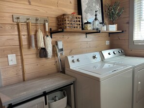 Un espace buanderie avec un lave-linge et un sèche-linge blancs, un lambris en bois, une étagère contenant des produits de nettoyage et un porte-linge suspendu avec des outils de nettoyage.