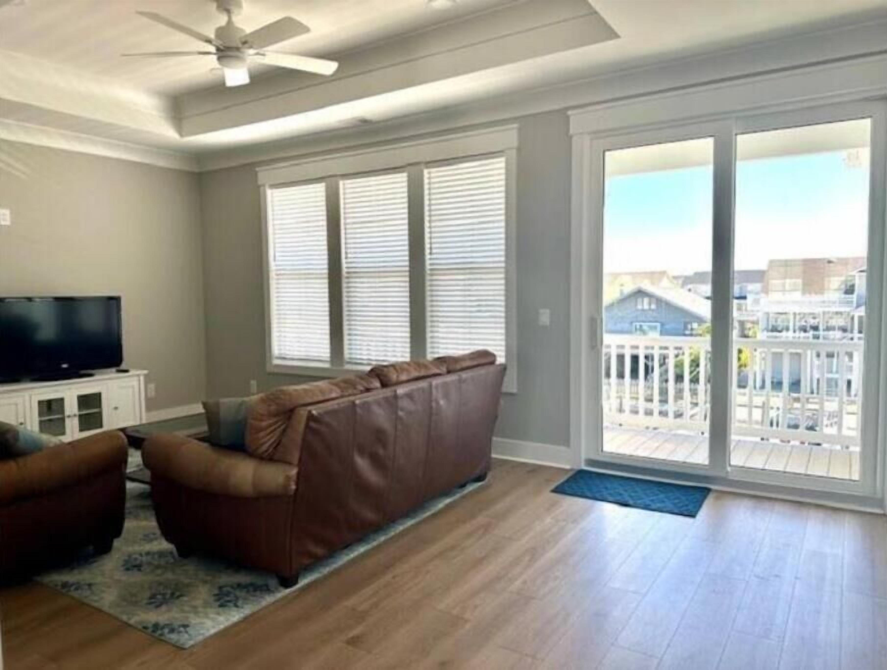 A living room with a brown leather sofa, a flat-screen TV on a white cabinet, and a large sliding glass door leading to a balcony.