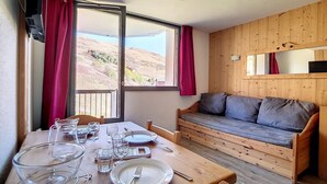 Un coin repas avec une table en bois dressée pour deux personnes, un canapé et une grande fenêtre donnant sur des collines.