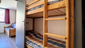 Une chambre avec un lit superposé, une structure en bois, une couvre-lit à carreaux et une fenêtre ornée d’un rideau rouge.