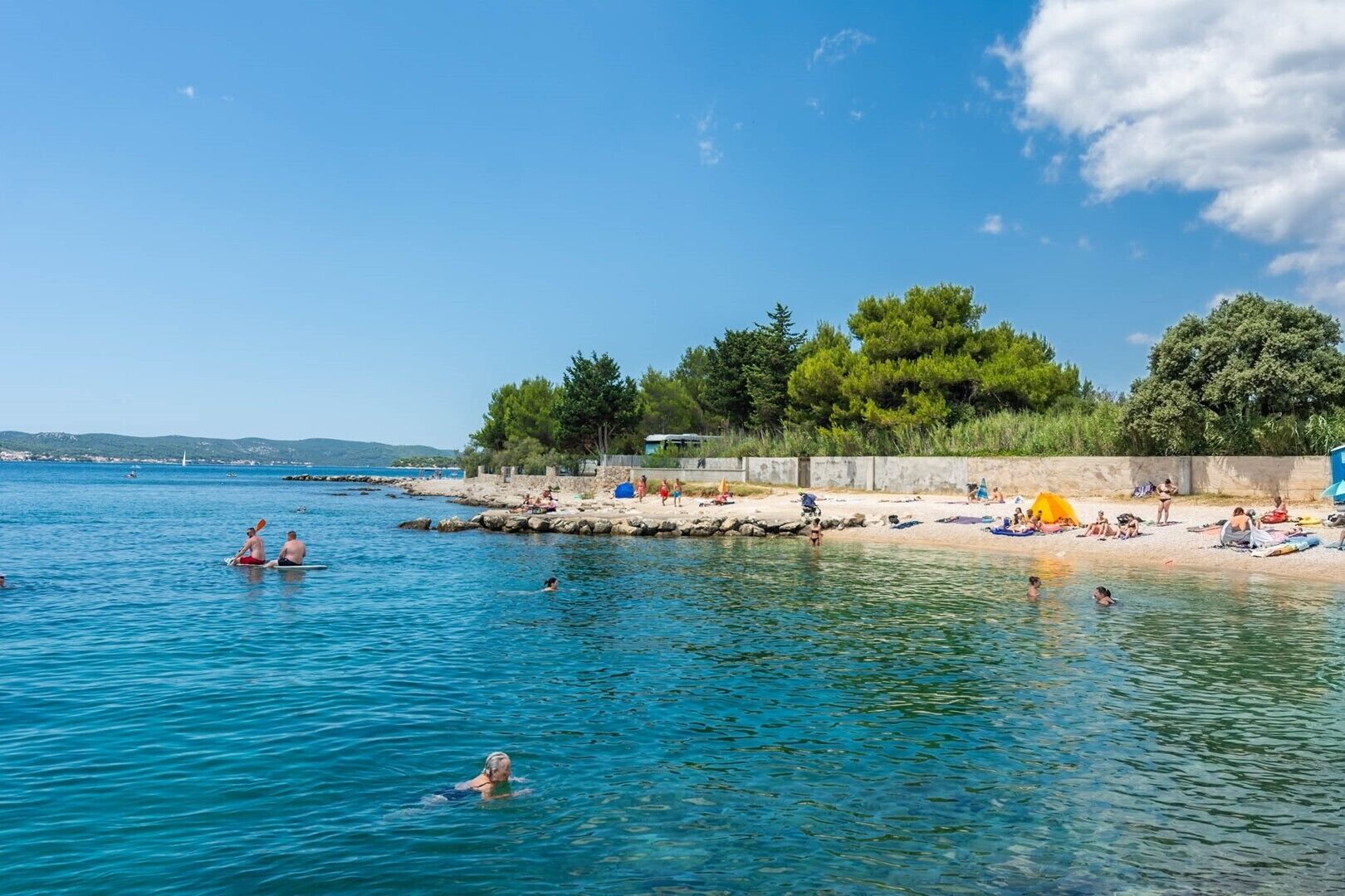 Ein Strand mit klarem, blauem Wasser, Menschen beim Schwimmen und Sonnenbaden, und eine bewaldete Gegend im Hintergrund.