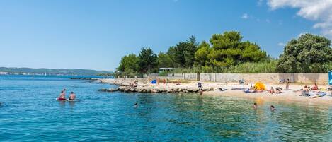 Ein Strand mit klarem, blauem Wasser, Menschen beim Schwimmen und Sonnenbaden, und eine bewaldete Gegend im Hintergrund.