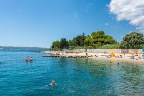 A beach with clear blue water, people swimming and sunbathing, and a forested area in the background.