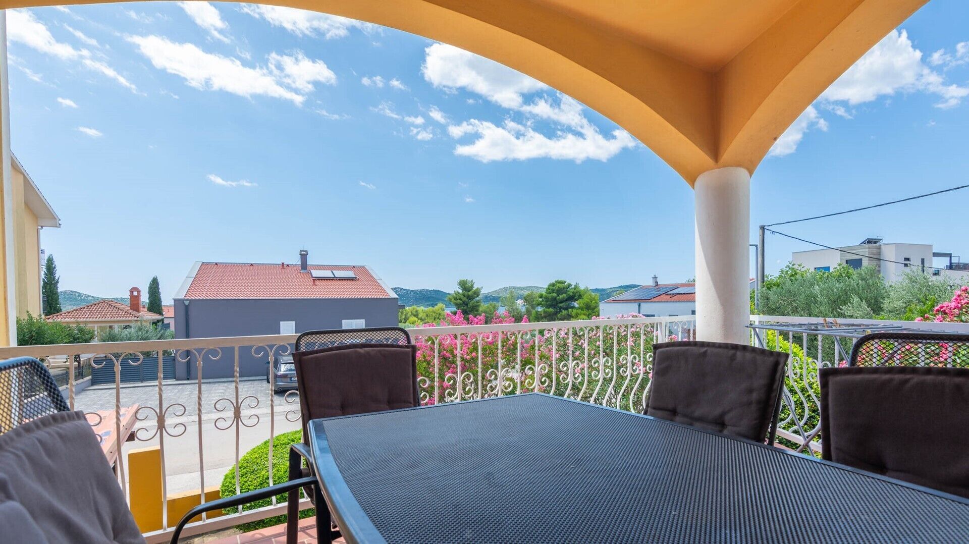 Een terras buiten met een tafel, stoelen, een balustrade en uitzicht op gebouwen en bomen.
