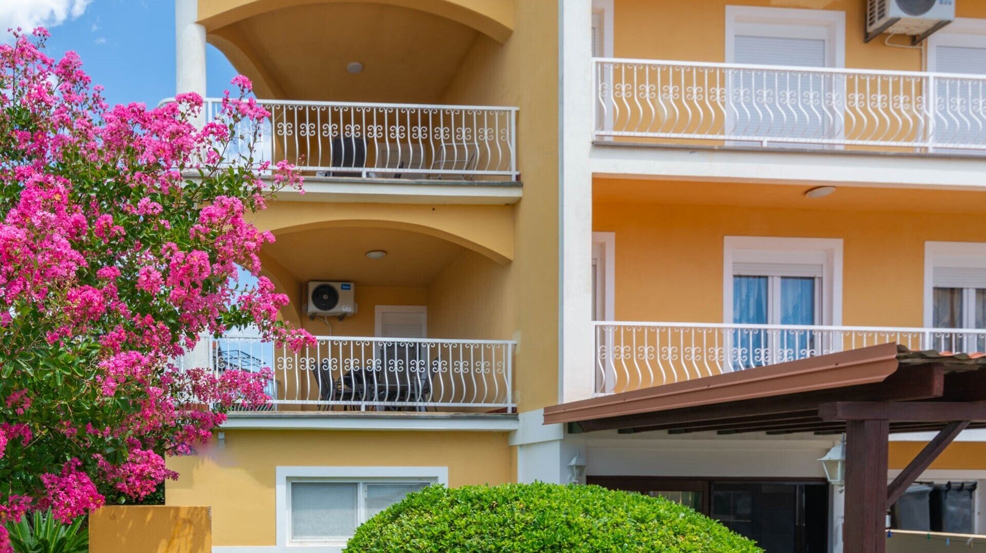 A multi-story building with balconies, air conditioning units, and a pink flowering tree.