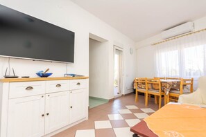 Una habitación con una televisión de pantalla plana montada en la pared, una mesa de comedor de madera con sillas, un armario blanco con cajones y un piso de baldosas.