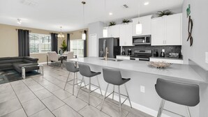 A modern kitchen with white cabinets, a central island with bar stools, and a dining area with a gray sofa and dining table. - Cozy 5 Bed Townhome with Splash Pool-9017DLD (Davenport)