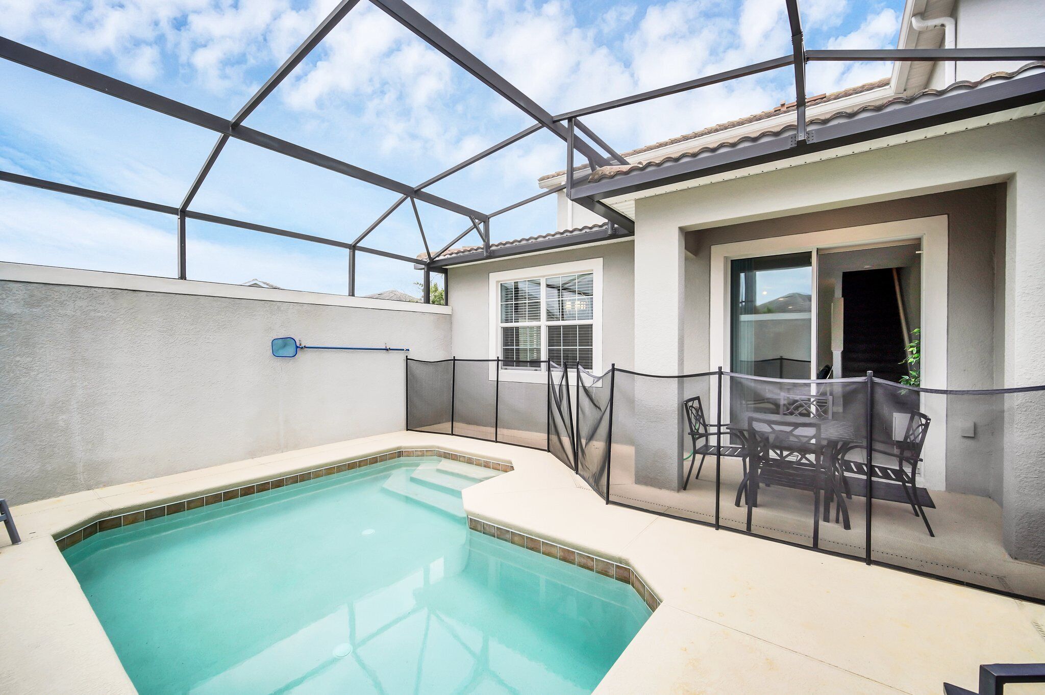 A covered swimming pool area with a patio table and chairs, a fenced enclosure, and a building with a tiled roof and windows.