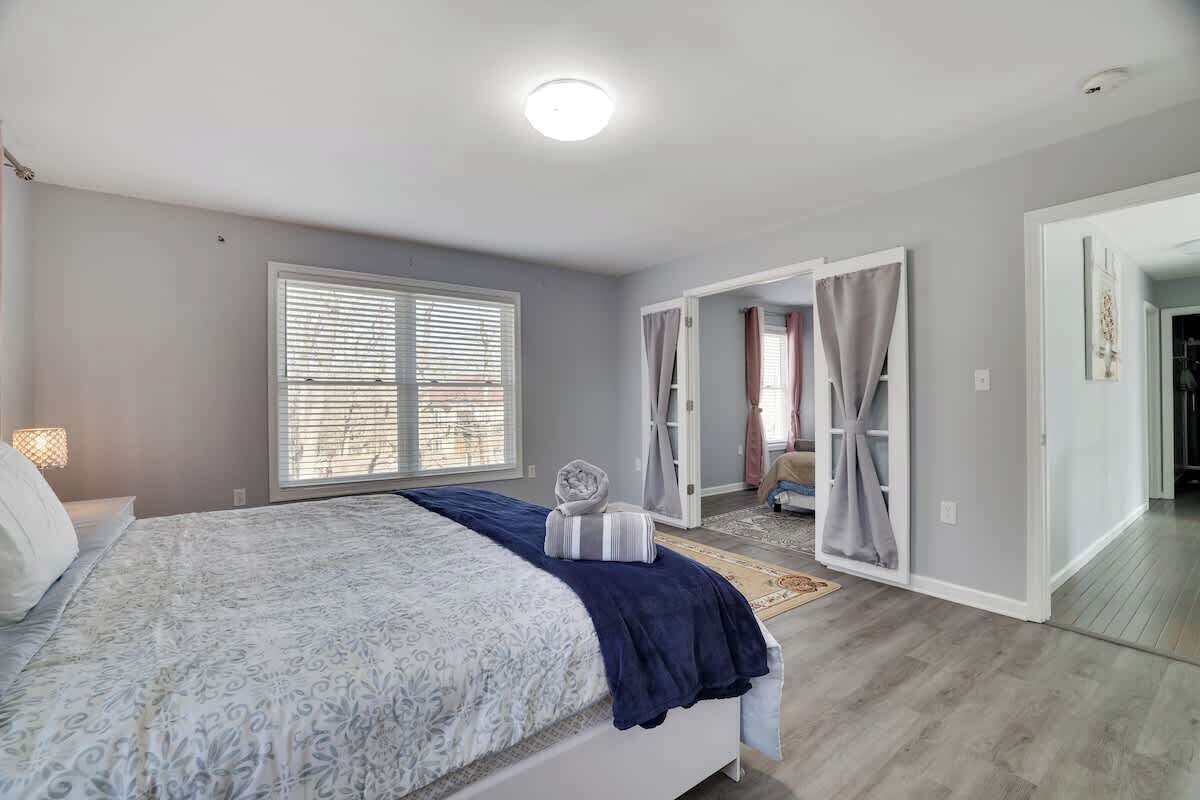 Une chambre avec un grand lit, une fenêtre avec des stores et une porte donnant sur une autre pièce.