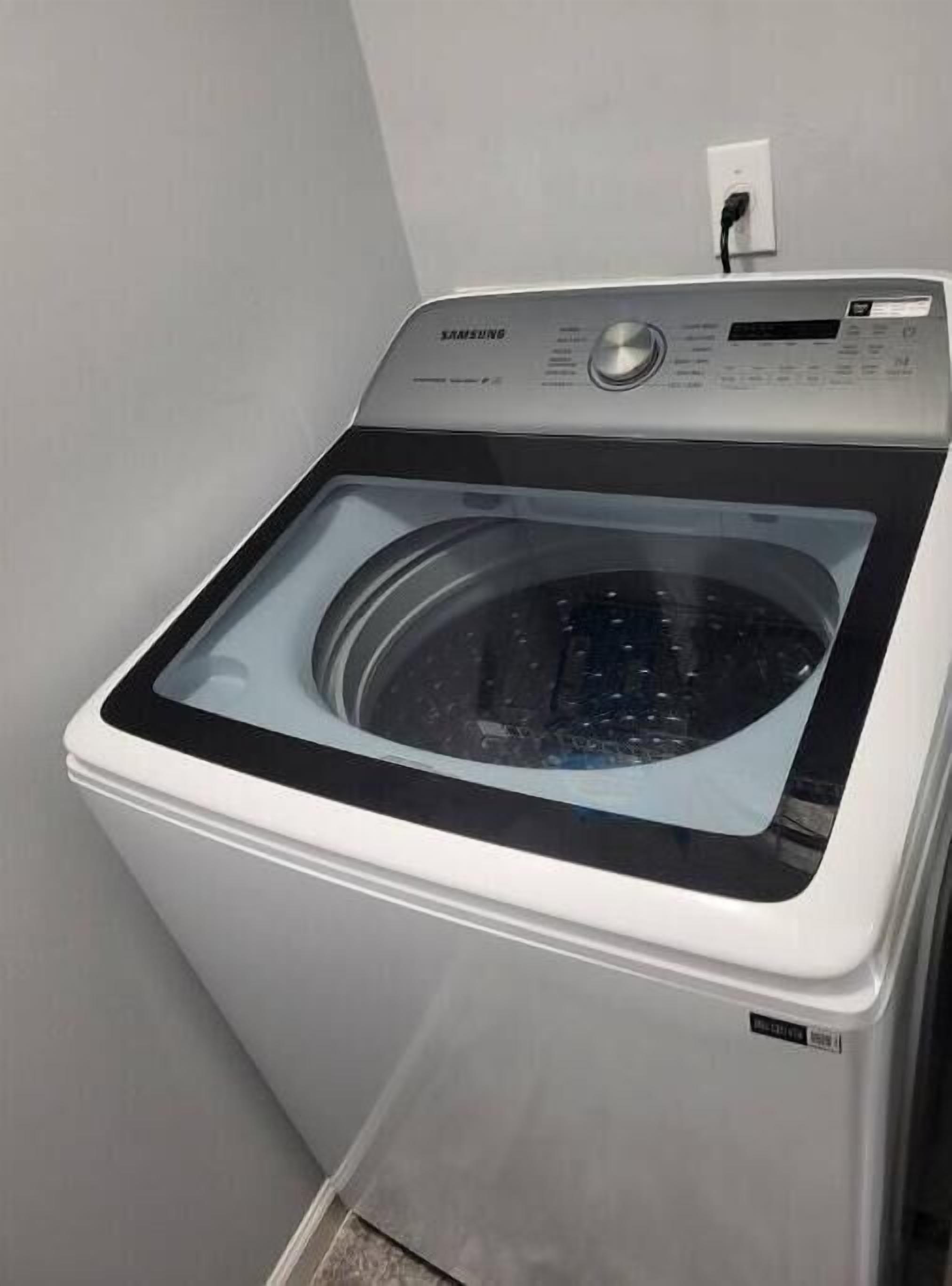 A Samsung front-loading washing machine in a laundry room with a white wall and a power outlet.