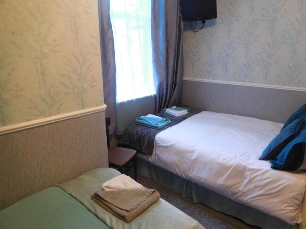 A small, single-room hotel room with two beds, a window with curtains, a flat-screen TV mounted on the wall, and a small table.