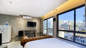 A modern hotel room with a large bed, a desk with a chair, a television, and a view of the cityscape through large windows. - Studio Vila Nova (São Paulo)