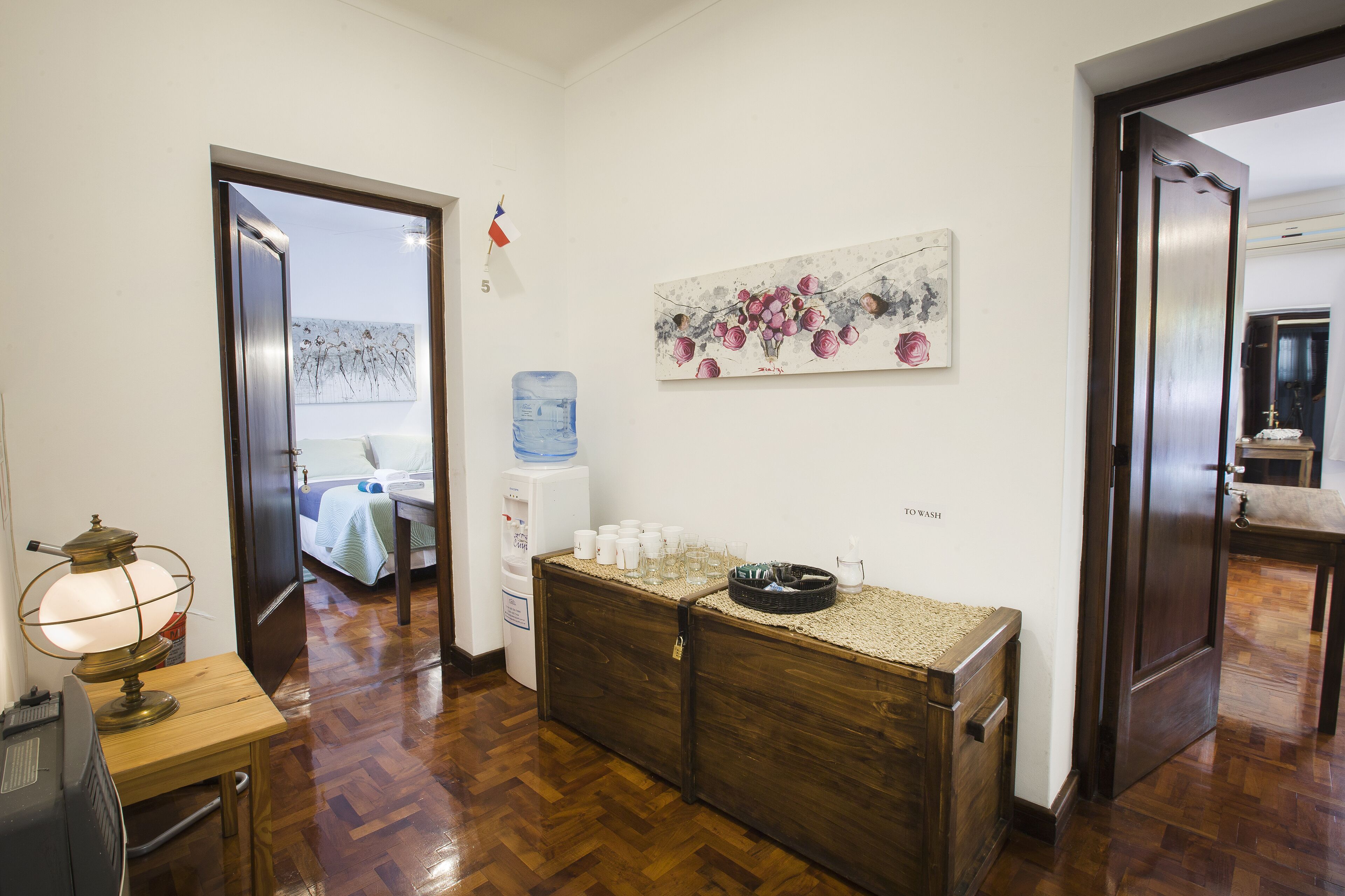 Vestíbulo de hotel con mostrador de recepción de madera, dispensador de agua, un cuadro en la pared y una cama visible a través de una puerta abierta.