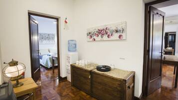 Vestíbulo de hotel con mostrador de recepción de madera, dispensador de agua, un cuadro en la pared y una cama visible a través de una puerta abierta.