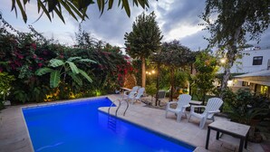 Une piscine rectangulaire avec un escalier, entourée de chaises longues blanches et d’une table, le tout se fondant dans un décor de verdure luxuriante et de plantes en fleurs.