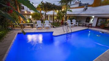 Une piscine à l’eau d’un bleu limpide, entourée de chaises et de tables blanches, et un bâtiment avec un toit en tuiles en arrière-plan.