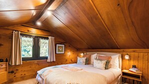 A cozy bedroom with a wooden ceiling, a bed with a quilt, a nightstand with books, and a window with curtains. - Le Petit Rêve - Lac de Montriond (Montriond)