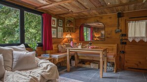 A cozy cabin interior with a sofa, a wooden dining table, a mirror, and a window with a red curtain. - Le Petit Rêve - Lac de Montriond (Montriond)