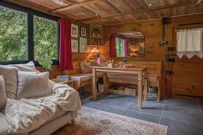 A cozy cabin interior with a sofa, a wooden dining table, a mirror, and a window with a red curtain.