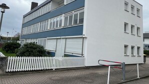 Um edifício de vários andares com fachada branca e detalhes em azul, apresentando uma cerca branca de piquete e uma entrada com portão.