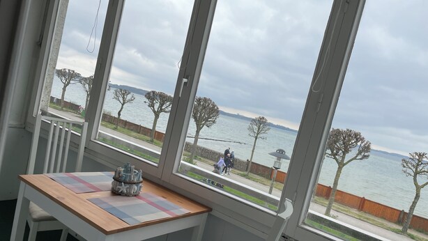 Quarto com uma grande janela com vista para a frente do mar. Há uma mesa de madeira com um jogo de toalha e um objeto decorativo.