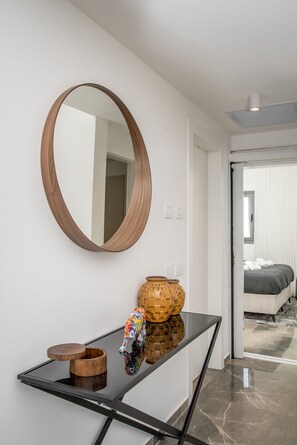 Ein modernes Interieur mit einem runden Spiegel, einem Konsolentisch mit Dekoration und einem Blick in ein Schlafzimmer mit Bett und Vorhängen.