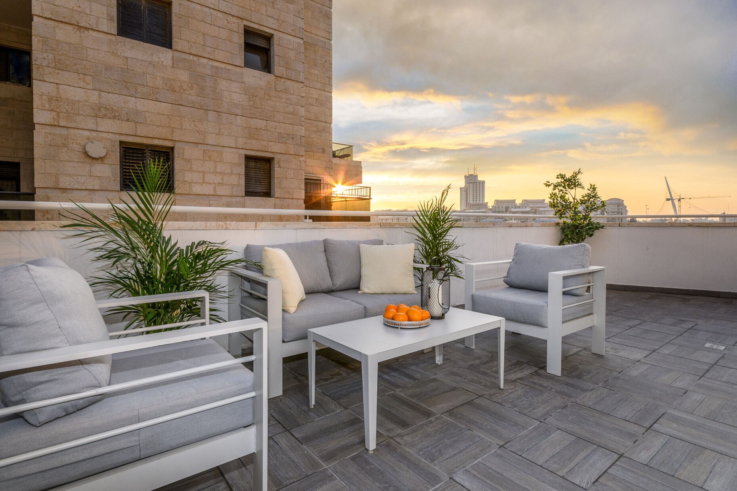 A rooftop terrace with a sofa set, a coffee table with oranges, and a view of the city skyline at sunset.